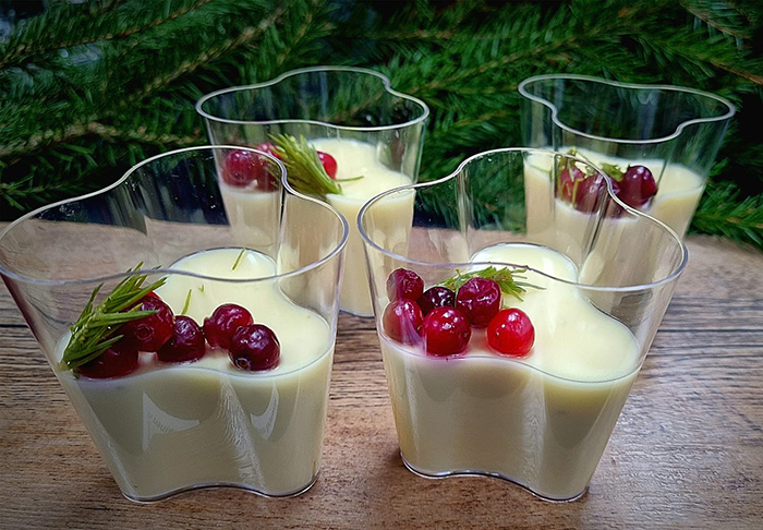 Neljässä lasimaljassa kuusenkerkkäposset-jälkiruokaa. Annosten päällä on karpaloita ja kuusenkerkkiä. Taustalla on koristeena kuusenoksia. 