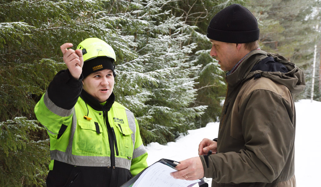 Metsäkoneenkuljettaja Mikko Inkinen ja Jukka Ruutiainen keskustelevat metsätiellä lehtometsän hoitohakkuusta. Taustalla on lumisia kuusia. 