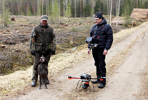 Tommi Kinnunen ja saksanseisoja Heta ja VideoDrone Oy:n Juhani Mikkola seisovat metsätiellä hakkuuaukon reunalla. Mikkolan kaulassa on lämpökamera ja maassa tiellä on drone. 