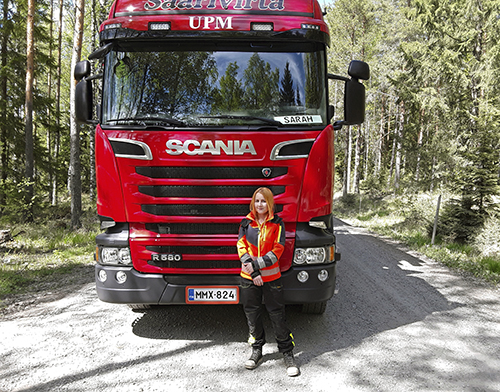 Puuauton kuljettaja Sarah Lindahl seisoo punaisen tukkirekan keulan edessä. Rekka on metsätiellä.