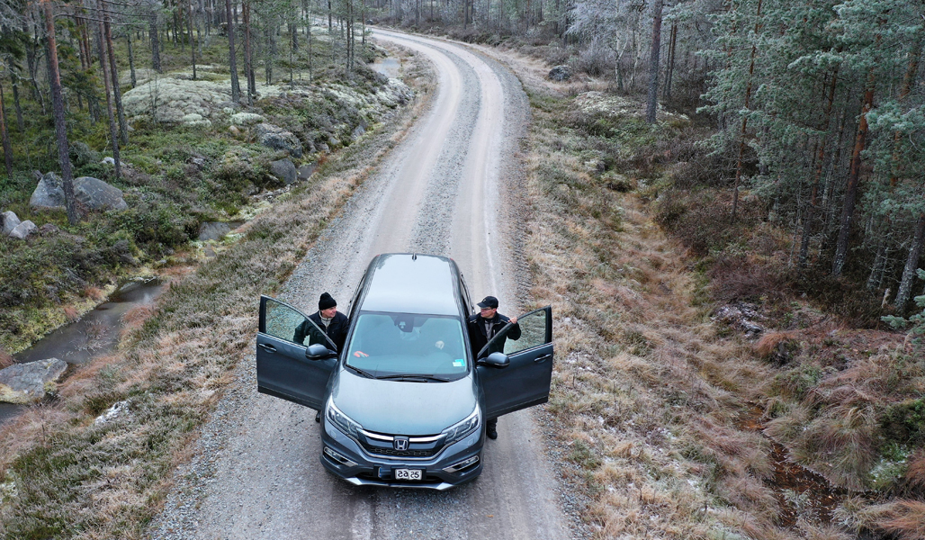 Kuvassa on auto kuvattuna ylhäältäpäin metsätiellä. Miehet nousevat autosta. 