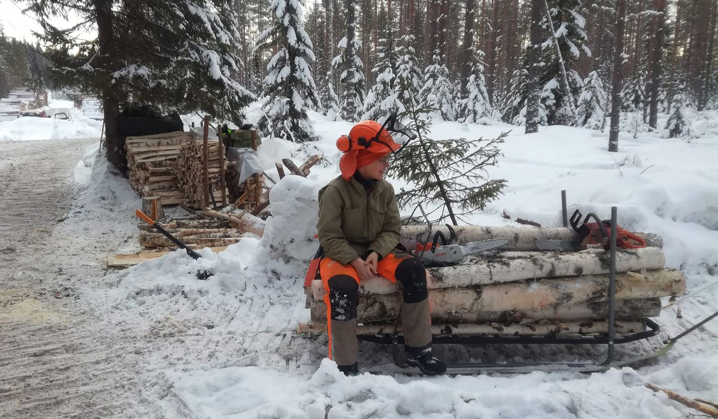 Pirjo Paldanius istuu metsurin suojavaatteissa tukkipinon päällä metsässä talvella. 