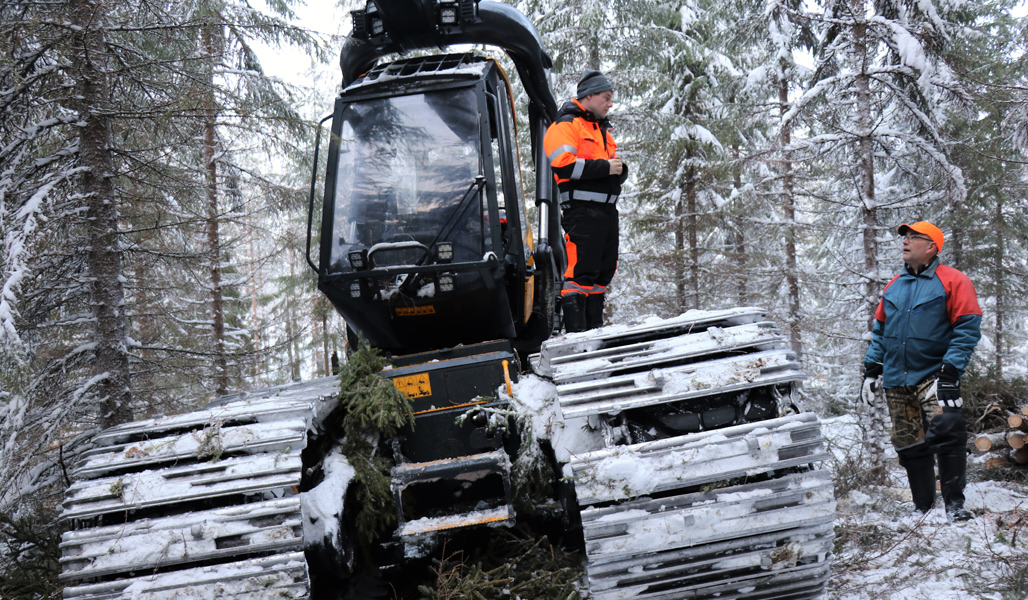 Metsäkoneenkuljettaja Mikko Ahonen seisoo metsäkoneen telojen päällä ja keskustelee Mikko Tiirolan kanssa. Ympärillä on lumista metsää. 