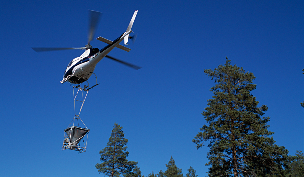 Helikopteri lentää kirkkaansinisellä taivaalla. Helikopterin alla olevasta astiasta pudotetaan tuhkaa metsään. Kuvan oikeassa alareunassa näkyy hieman männynlatvaa. 