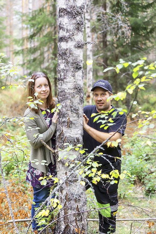 Paula Viilo ja Harri Viilo kurkistavat koivun rungon takaa. Taustalla on metsää.