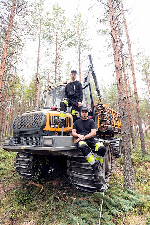 Viilon perheen vanhin poika Lauri Viiloon noussut seisomaan metsätraktorin päälle. Traktorin perävaunuun on kuormattu puupöllejä. Isä Harri Viilo nojaa metsätraktoriin. 